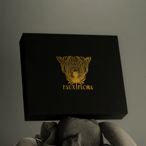 Black box with gold Faux Flora logo and text held by a rocks against a gray background