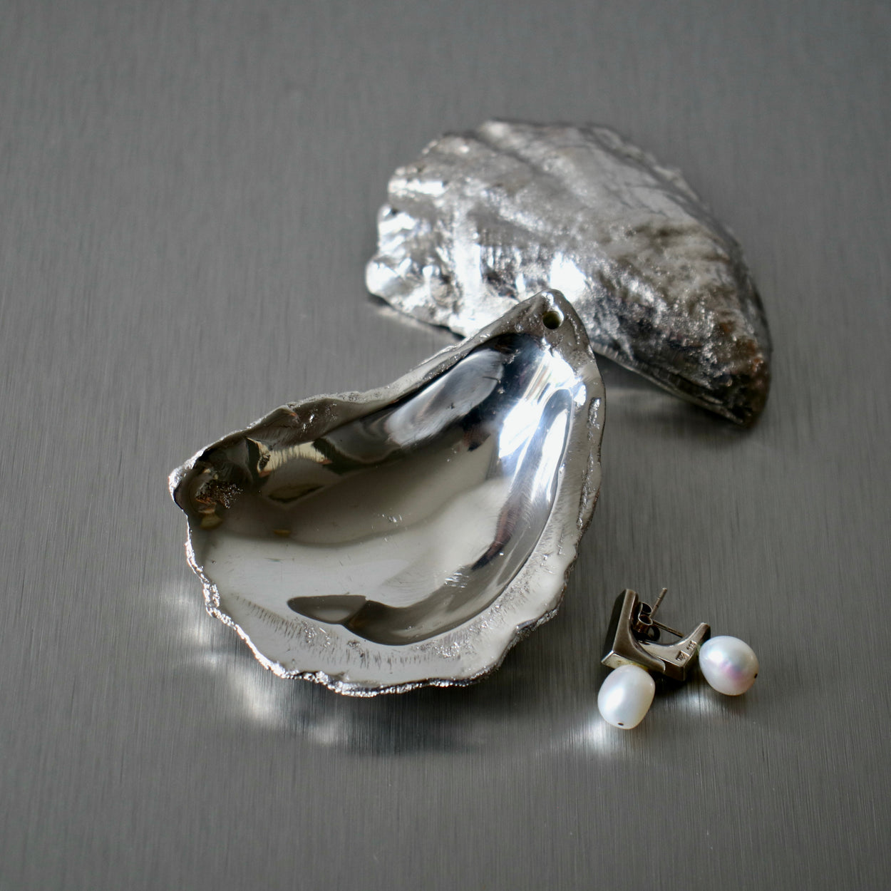 Silver oyster shell incense holders with pearl earrings on a stainless steel bench top.