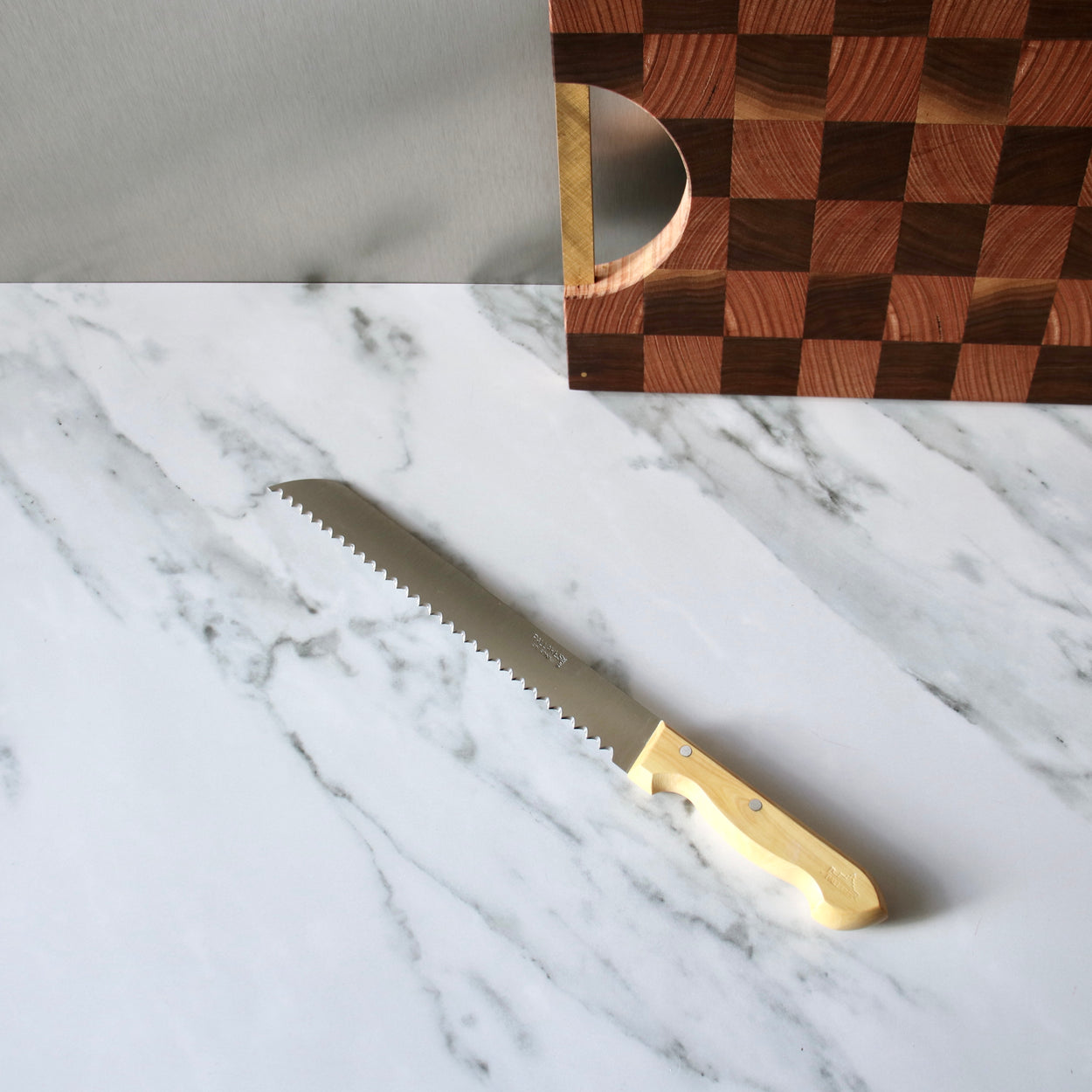 Pallares Solsona 25cm Box Wood Stainless Steel Bread Knife on marble bench with chopping board in background.