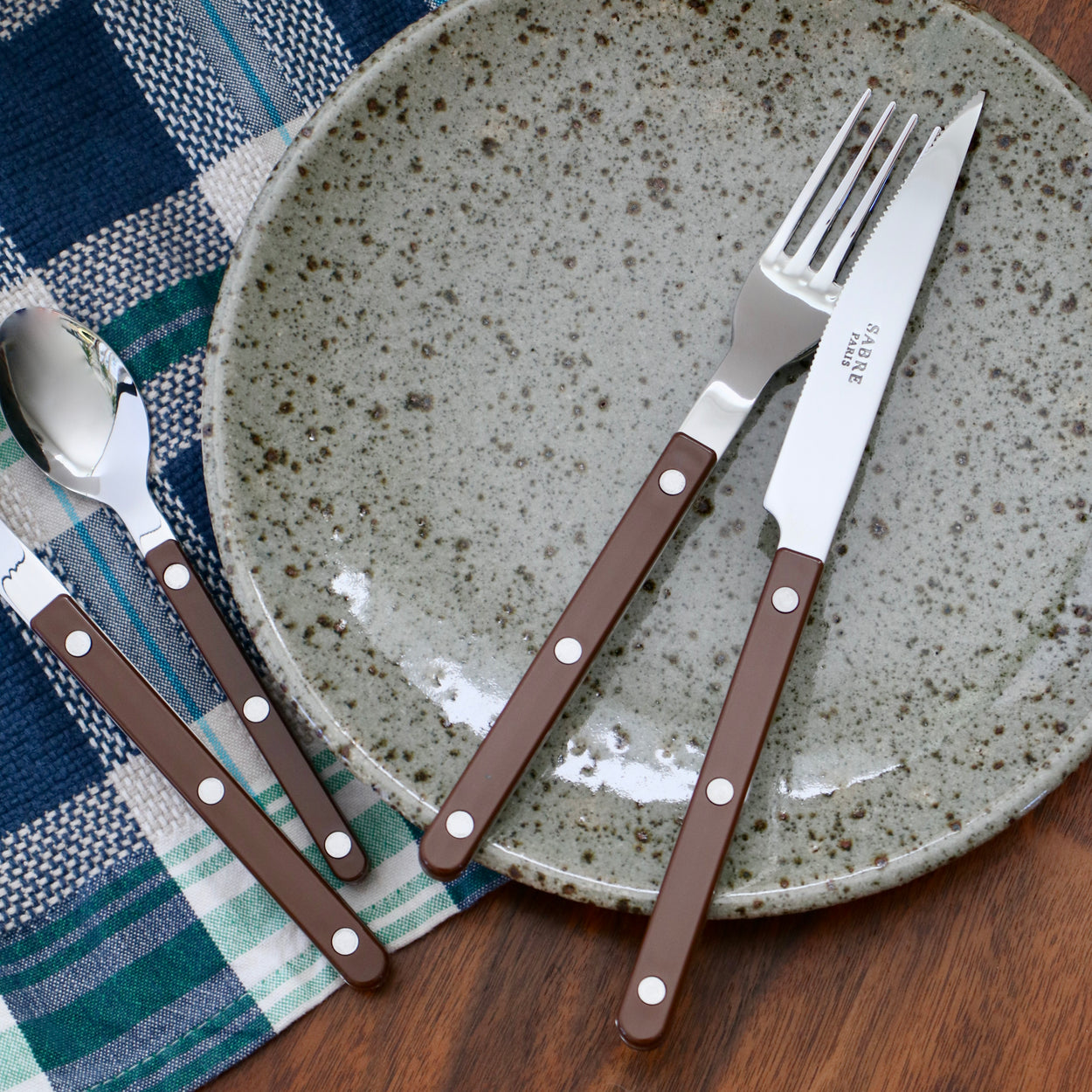 Sabre Paris Bistrot Chocolate Brown Cutlery 4 Piece Set on ceramic plate with blue napkin and wood table.