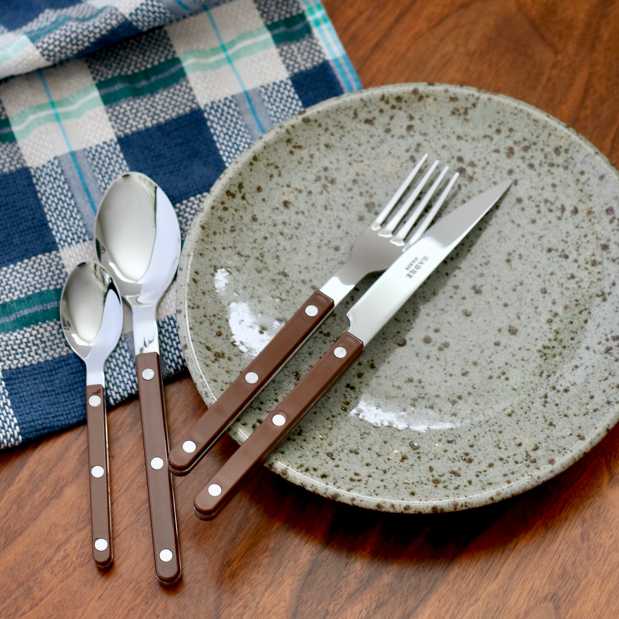 Sabre Paris Bistrot Chocolate Brown Cutlery 4 Piece Set on ceramic plate with blue napkin and wood table.