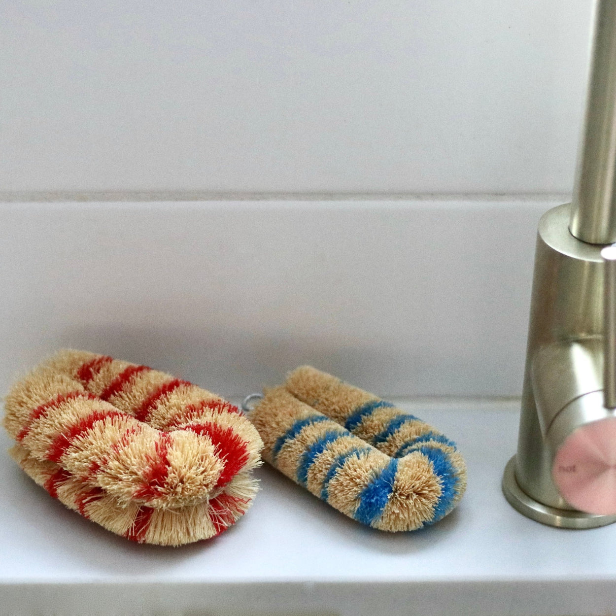 Two Japanese Striped Tawashi pot scrub brushes on white tile shelf with gold tapware.