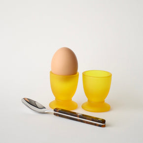 Two yellow egg cups with a brown egg and a silver spoon on a white background