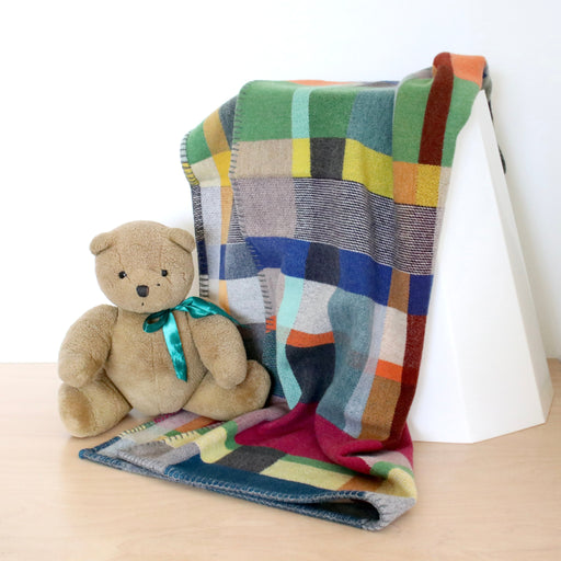 Colourful wool blanket draped over a white surface with a teddy bear sitting on it.