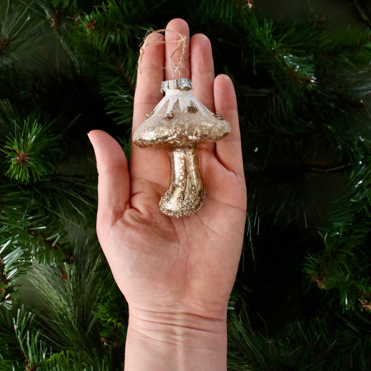 Hand holding a glittery mushroom ornament against a green pine tree background