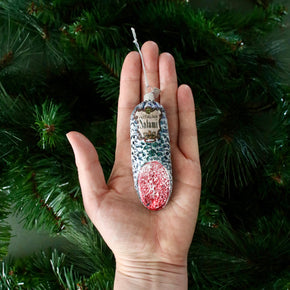 Hand holding a decorative ornament shaped like a salami with red and silver design against a green Christmas tree background.
