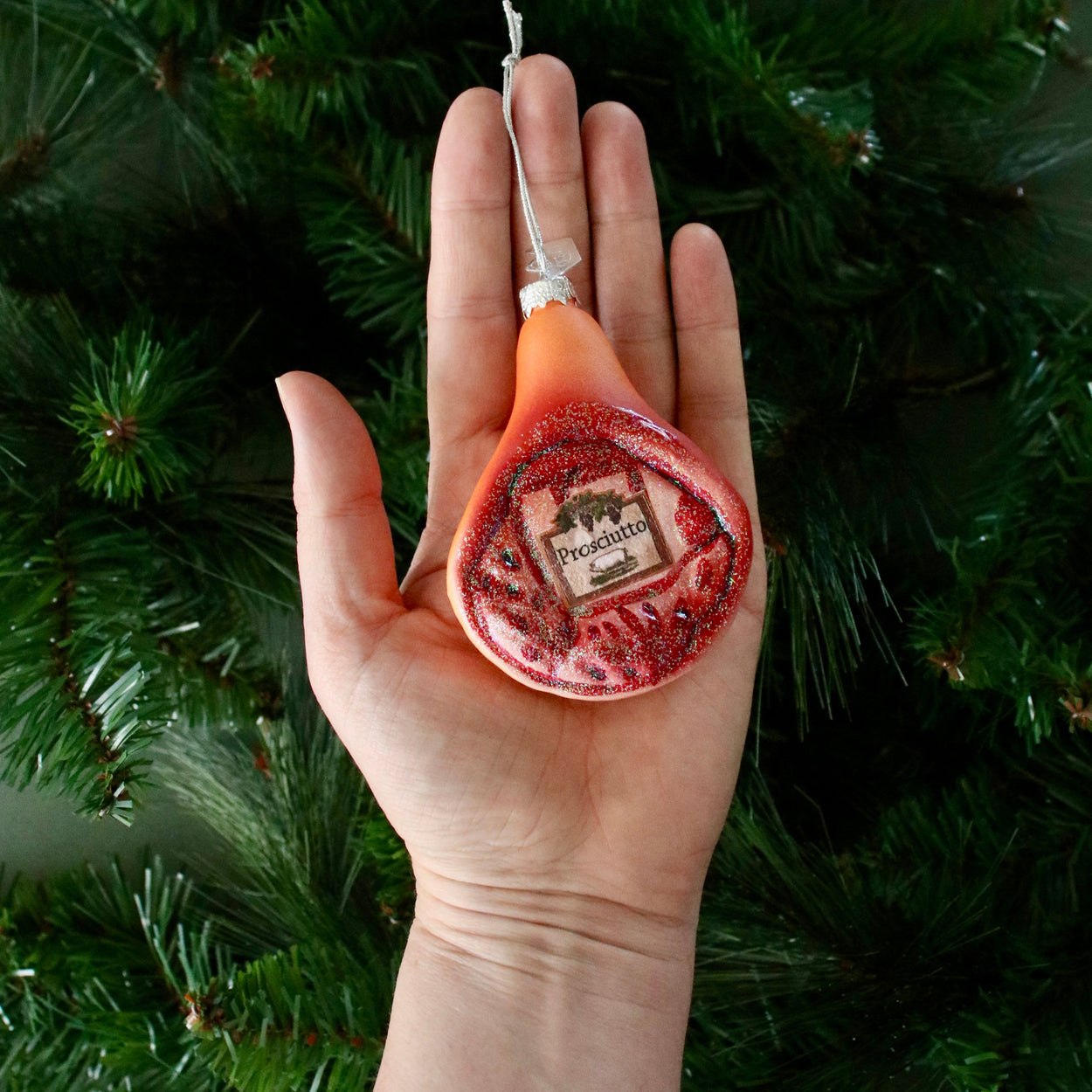 Hand holding a Ham shaped ornament against a green Christmas tree background