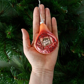 Hand holding a Ham shaped ornament against a green Christmas tree background