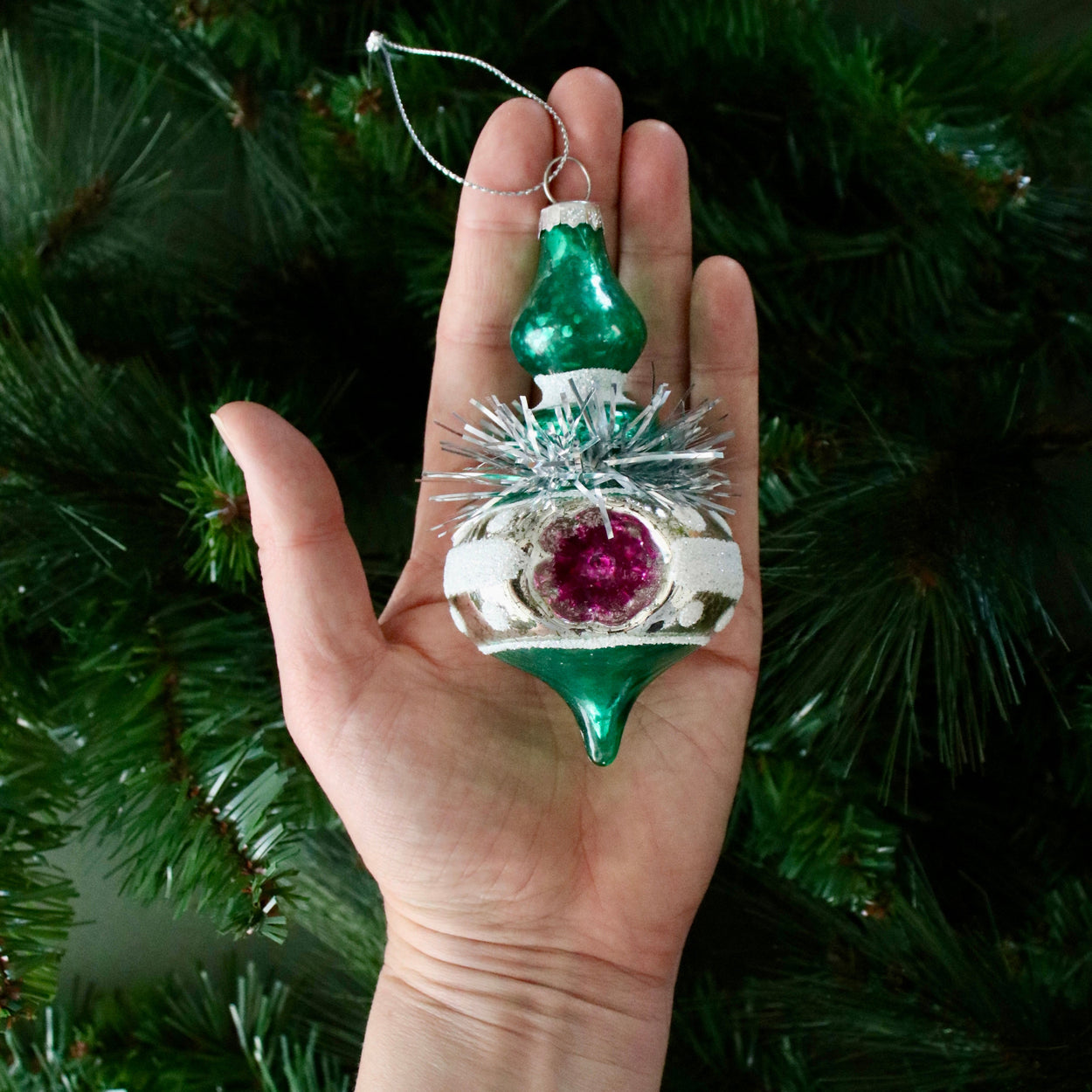 Hand holding a decorative Christmas ornament with a green top and purple center against a green tree background