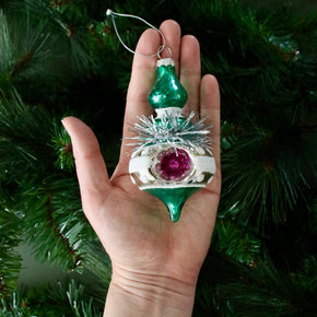 Hand holding a decorative Christmas ornament with a green top and purple center against a green tree background