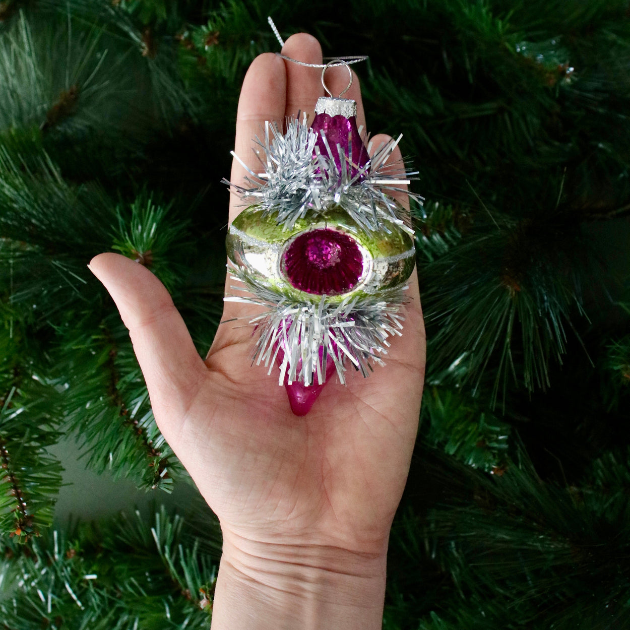 Hand holding a decorative Christmas ornament with green, purple, and silver colors against a green Christmas tree background.