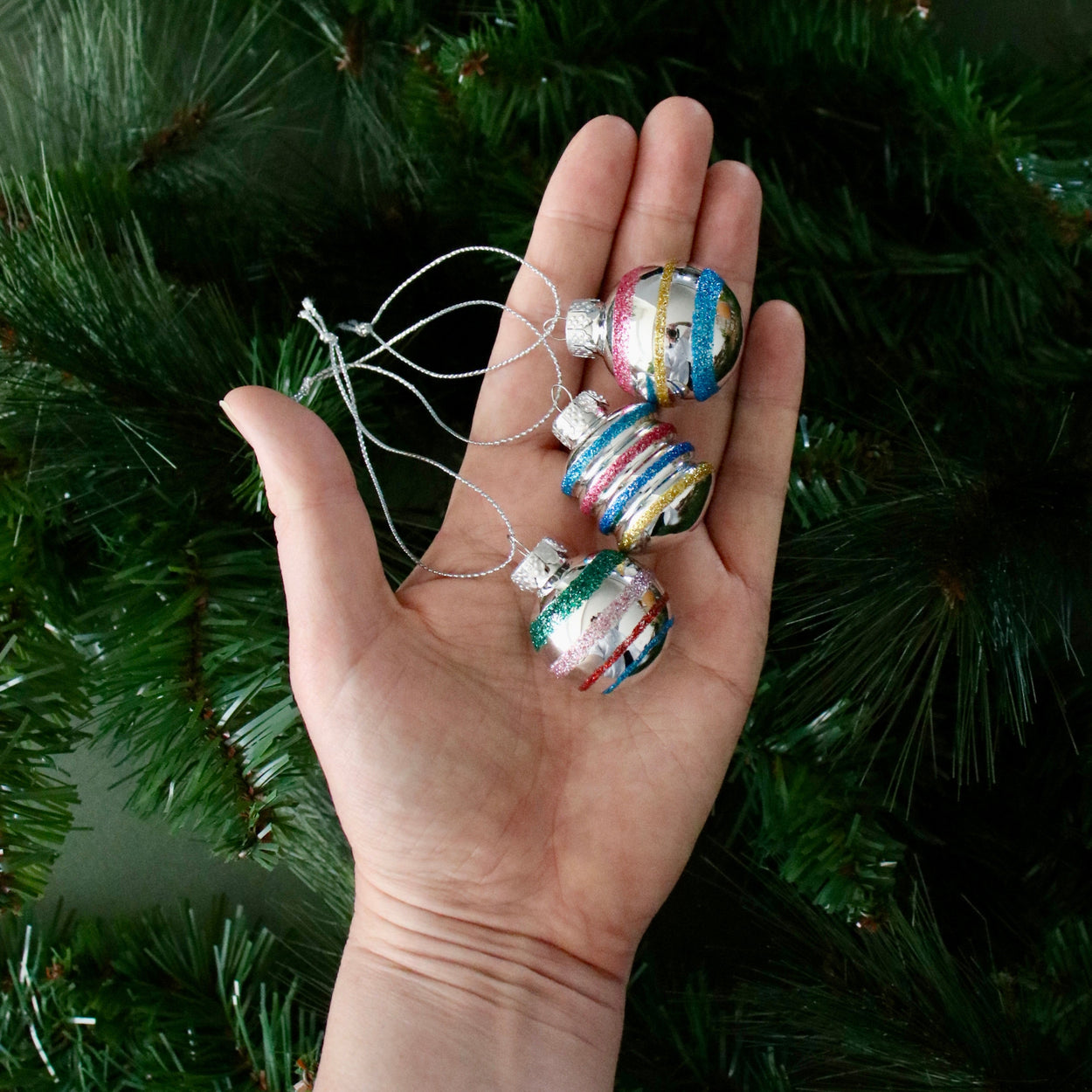 Hand holding three colorful glass Christmas ornaments against a green tree background
