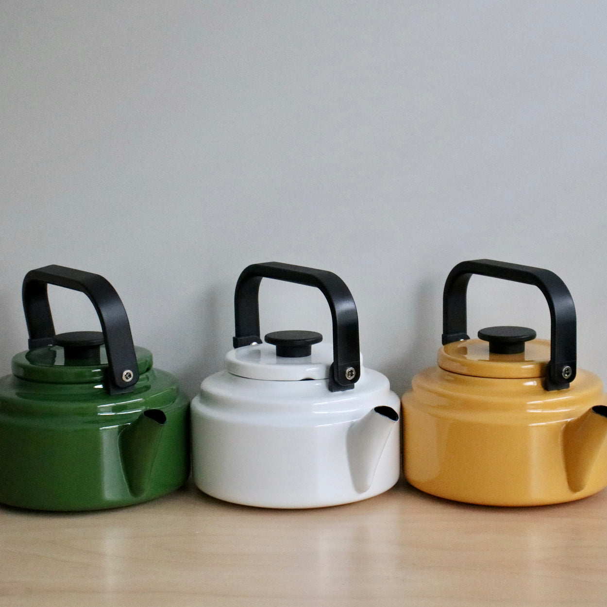 Three colorful kettles (green, white, yellow) with black handles on a wooden surface.