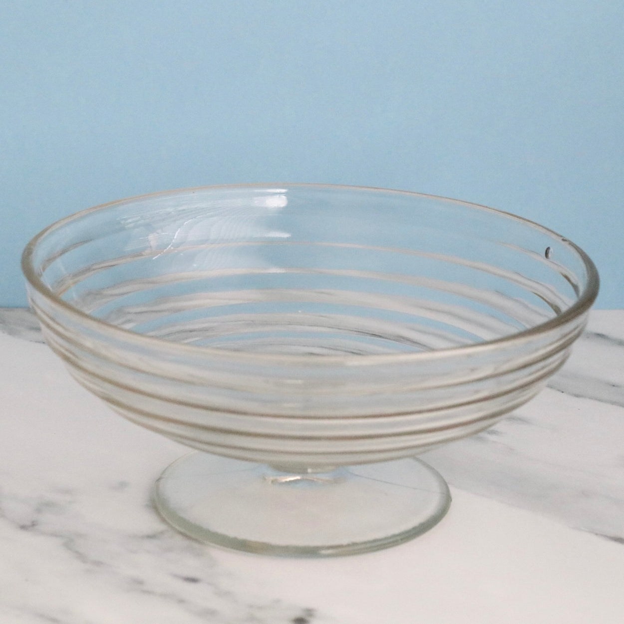 Clear glass footed bowl on a marble surface with a light blue background