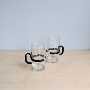 Two clear glass mugs with black handles on a beige surface.