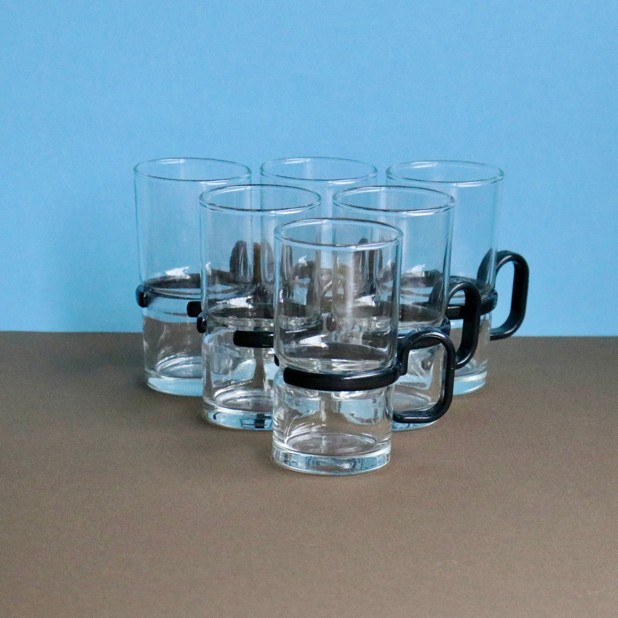 Set of clear glass mugs with black handles on a brown surface with a blue background