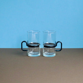 Two clear glass mugs with black handles on a brown surface and blue background