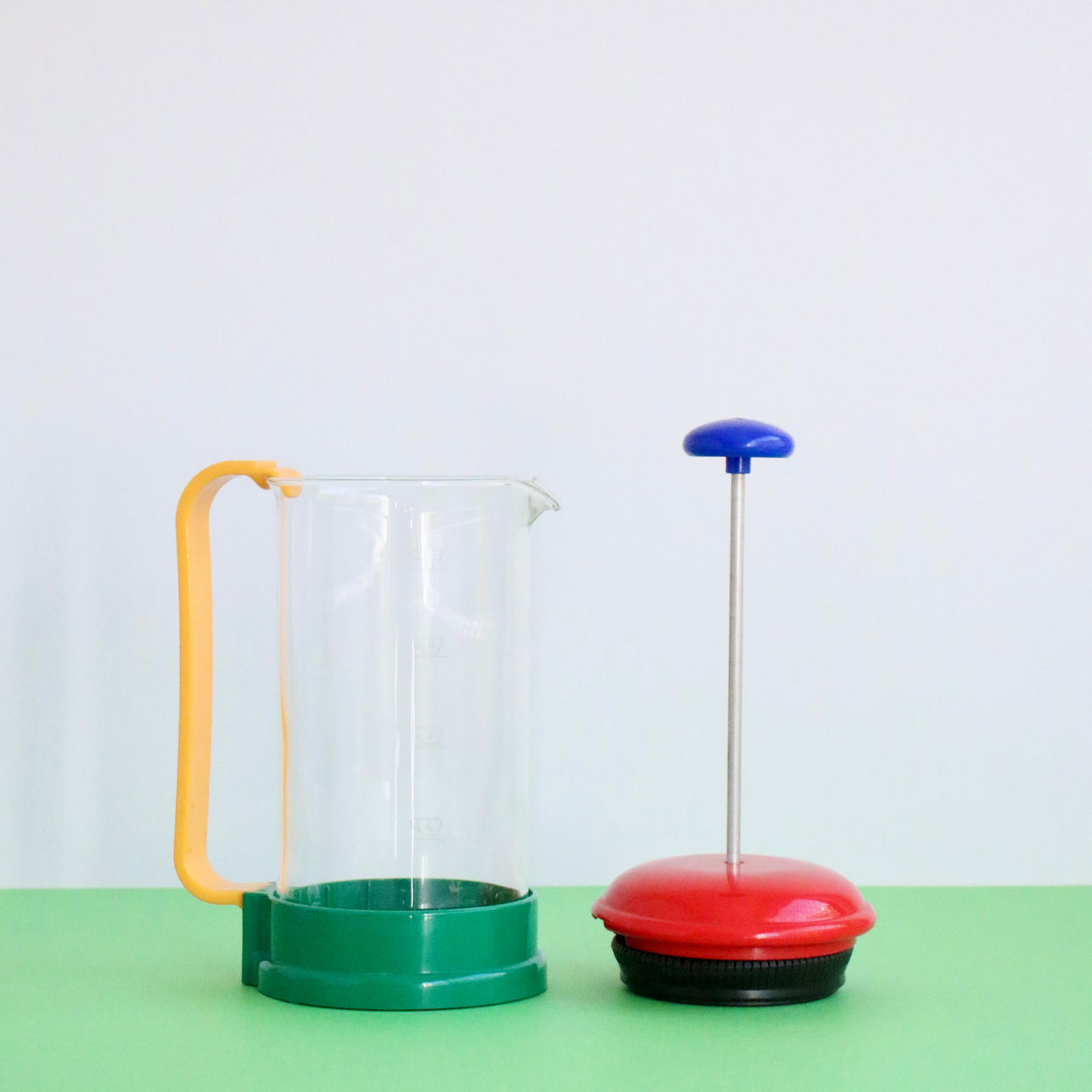 French Press with yellow handle and green base, and red and blue lid on a light green surface.