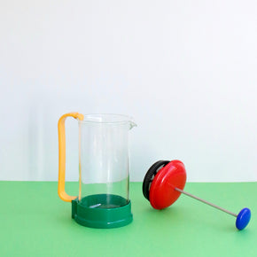 French Press with yellow handle and red and black lid on a green surface