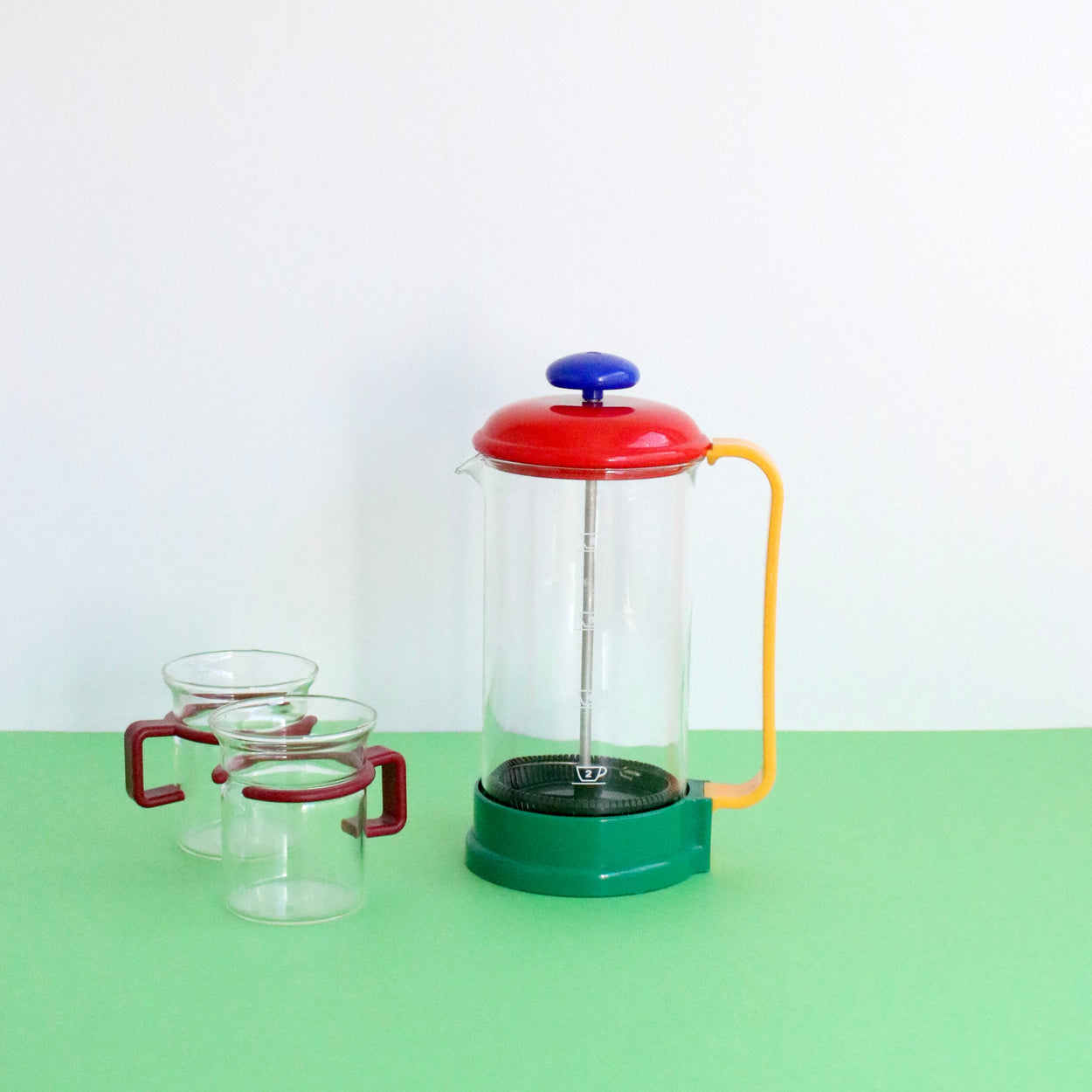 Colorful French press coffee maker with two glass mugs on a green surface.