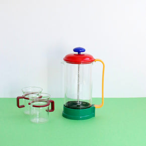 Colorful French press coffee maker with two glass mugs on a green surface.
