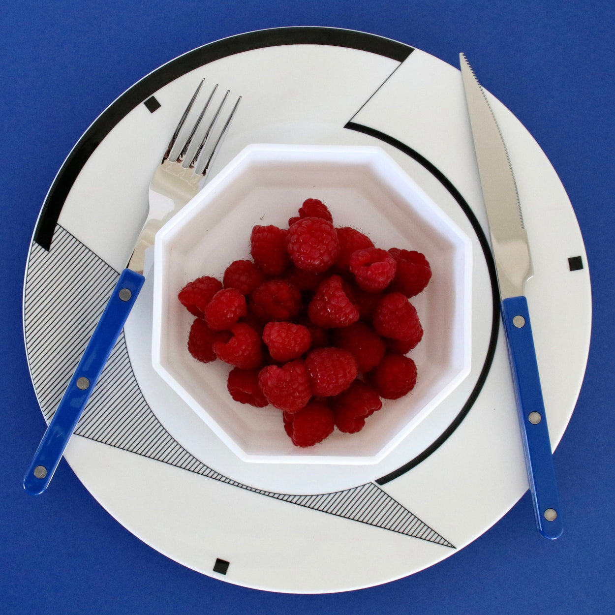 White bowl with raspberries on a decorative plate with cutlery against a blue background
