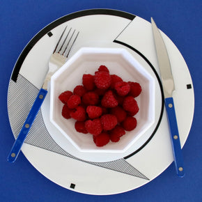 White bowl with raspberries on a decorative plate with cutlery against a blue background