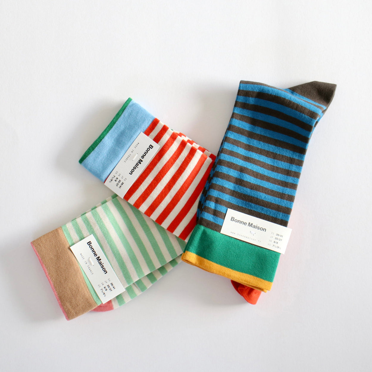 Three pairs of striped socks from Bonne Maison arranged on a plain white background. The socks feature various stripe patterns including blue and black, red and white, and green and white, with one pair showing a brown and orange base.