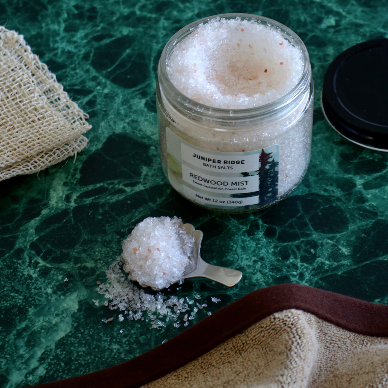 A clear glass jar of Juniper Ridge Redwood Mist bath salts sits on a green marble surface, with a small metal scoop holding some of the white salt crystals in front. A beige burlap cloth and a dark lid are partially visible around the scene.