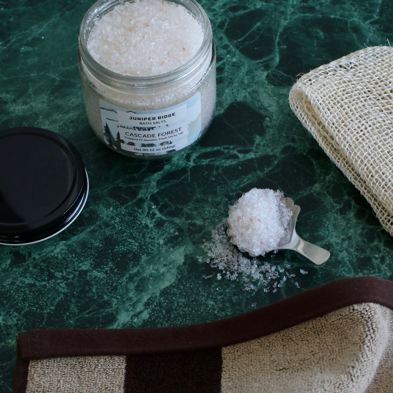 A glass jar of Juniper Ridge Apothecary Cascade Forest Bath Salt with its black lid and a metal scoop filled with the white salt crystals, arranged on a green marble surface next to a beige burlap cloth.