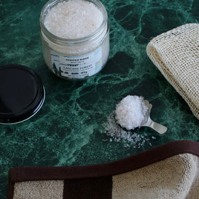 A glass jar of Juniper Ridge Apothecary Cascade Forest Bath Salt with its black lid and a metal scoop filled with the white salt crystals, arranged on a green marble surface next to a beige burlap cloth.