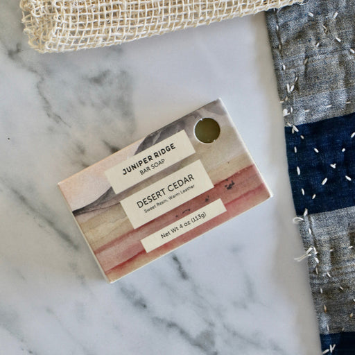 A bar of soap labeled 'JUNPER RIDGE BAR SOAP DESERT CEDAR' placed on a white marble surface, with a woven beige cloth at the top and a blue-gray patchwork textile at the right edge.