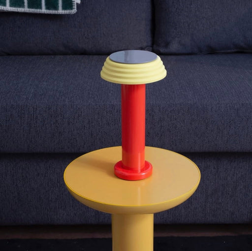 Colorful cylindrical Sowden PL1 lamp with a red base and yellow top against a dark blue sofa.