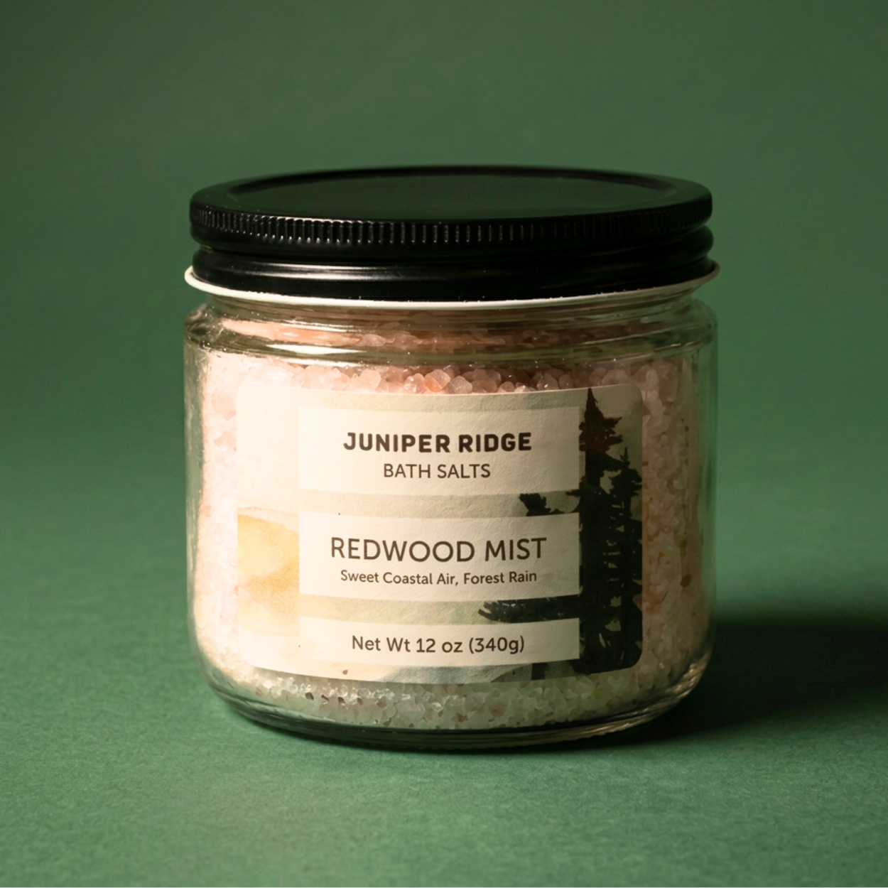 A glass jar of Juniper Ridge Bath Salts, Redwood Mist, filled with pinkish salt crystals and sealed with a black lid, set against a dark green background