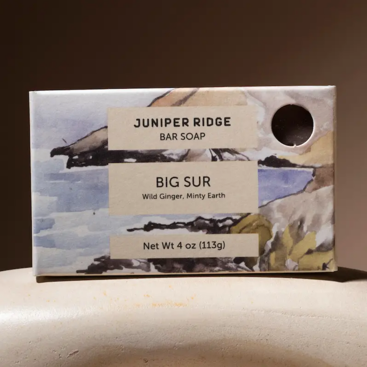 A rectangular bar of Juniper Ridge Big Sur soap in a watercolor-style box with beige labels and a circular cutout, photographed on a light surface against a dark brown background.