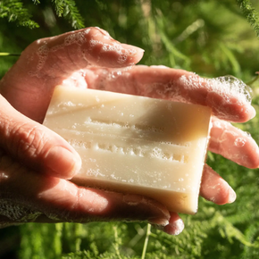 A handmade rectangular bar of soap in a creamy beige color, held in hands covered in lathered soap against a blurred green outdoor background.