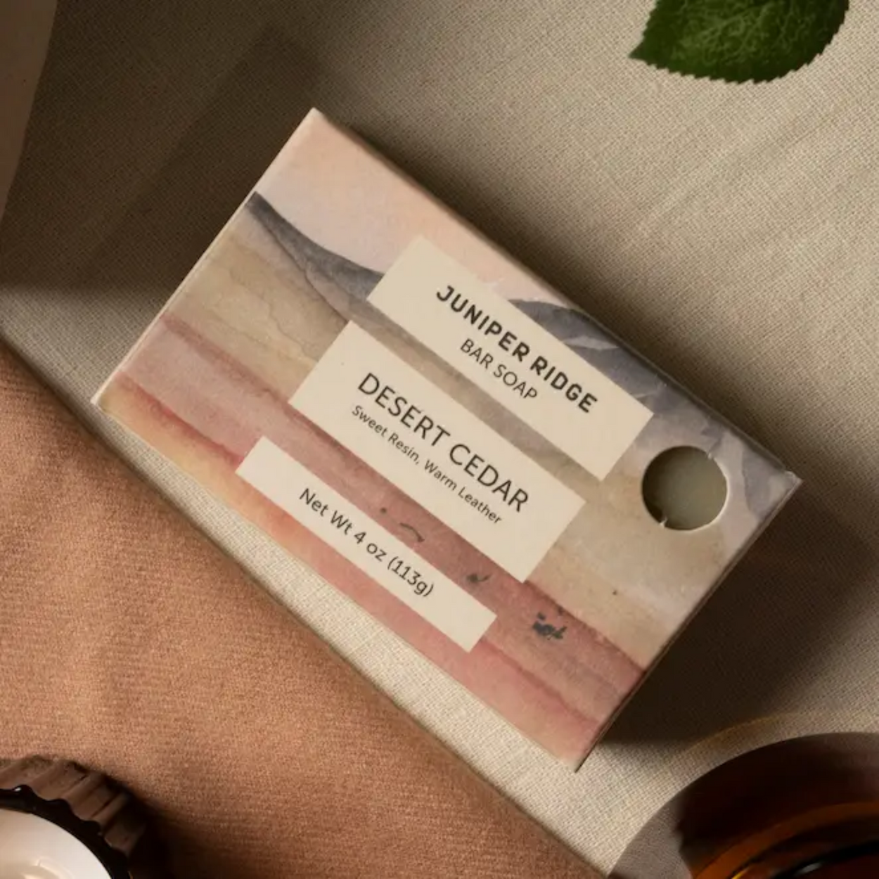 A rectangular bar soap box labeled 'Juniper Ridge Bar Soap' and 'Desert Cedar' lies on a beige fabric surface with a soft neutral background and a small green leaf in the corner.