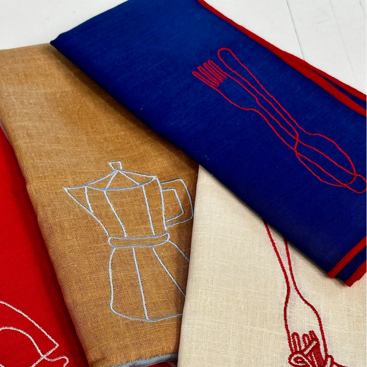 Four folded linen napkins in red, tan, blue, and cream, each featuring a minimalist embroidered kitchen utensil or food outline in contrasting thread.