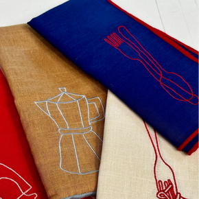 Four folded linen napkins in red, tan, blue, and cream, each featuring a minimalist embroidered kitchen utensil or food outline in contrasting thread.