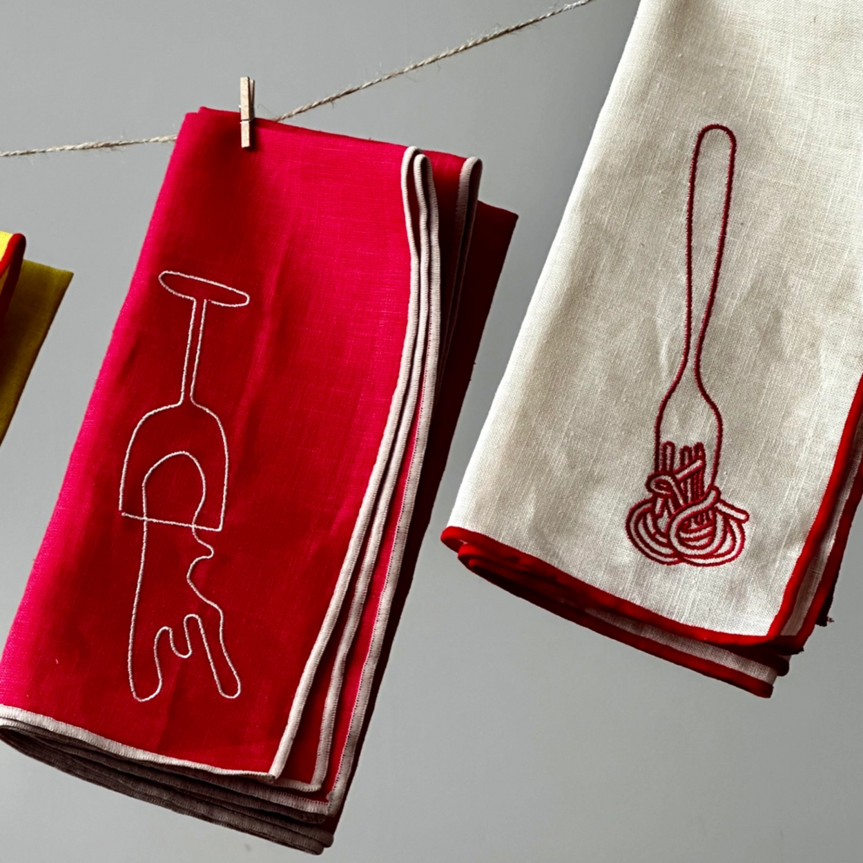 Two folded linen napkins hanging from a string with wooden clothespins against a neutral gray background. The left napkin is bright red with a white outline of a wine glass, and the right napkin is off-white with a dark red outline of spaghetti and a fork.