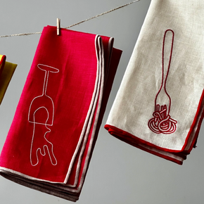 Two folded linen napkins hanging from a string with wooden clothespins against a neutral gray background. The left napkin is bright red with a white outline of a wine glass, and the right napkin is off-white with a dark red outline of spaghetti and a fork.