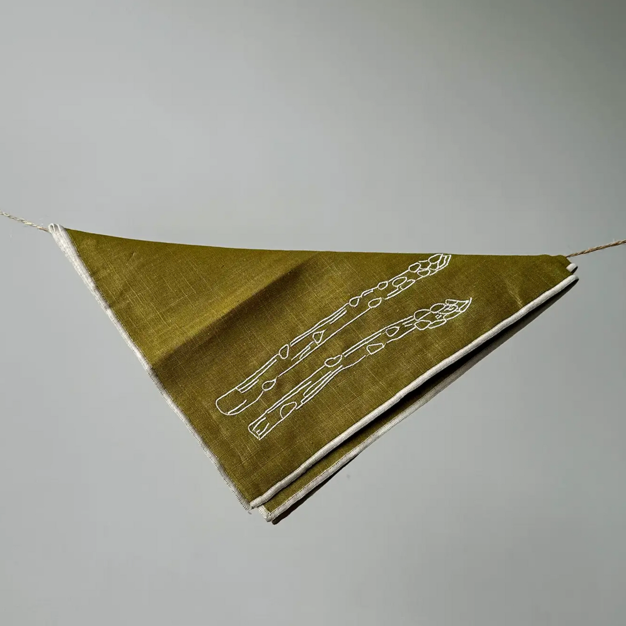 A triangular fabric napkin in olive green with white embroidered asparagus motifs, suspended by strings against a neutral gray background.