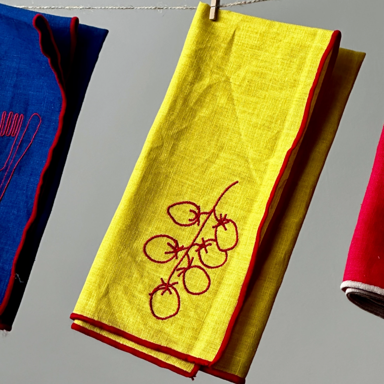 A folded yellow napkin with red embroidered fruit motifs and a red border hangs from a clothesline with a wooden clip, flanked by other colorful napkins against a neutral gray background.
