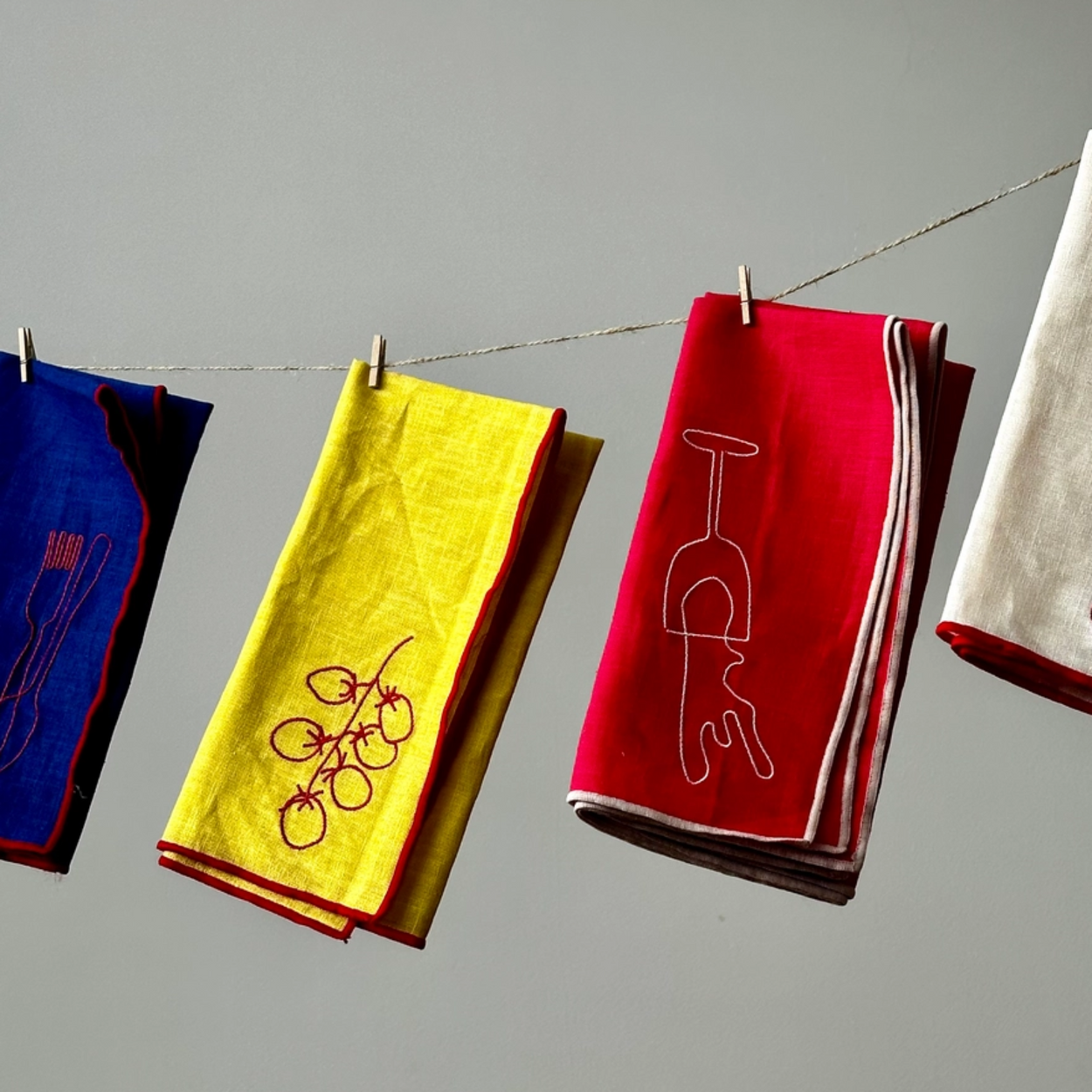 Four folded napkins in blue, yellow, red, and white hang from a string with wooden clothespins against a plain gray wall. Each napkin has a simple red line-art illustration of a kitchen utensil or object.
