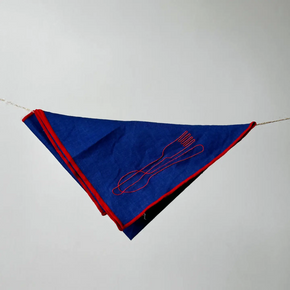 A blue triangular napkin with red trim and a red embroidered utensil design, hanging from a string against a plain light gray background.