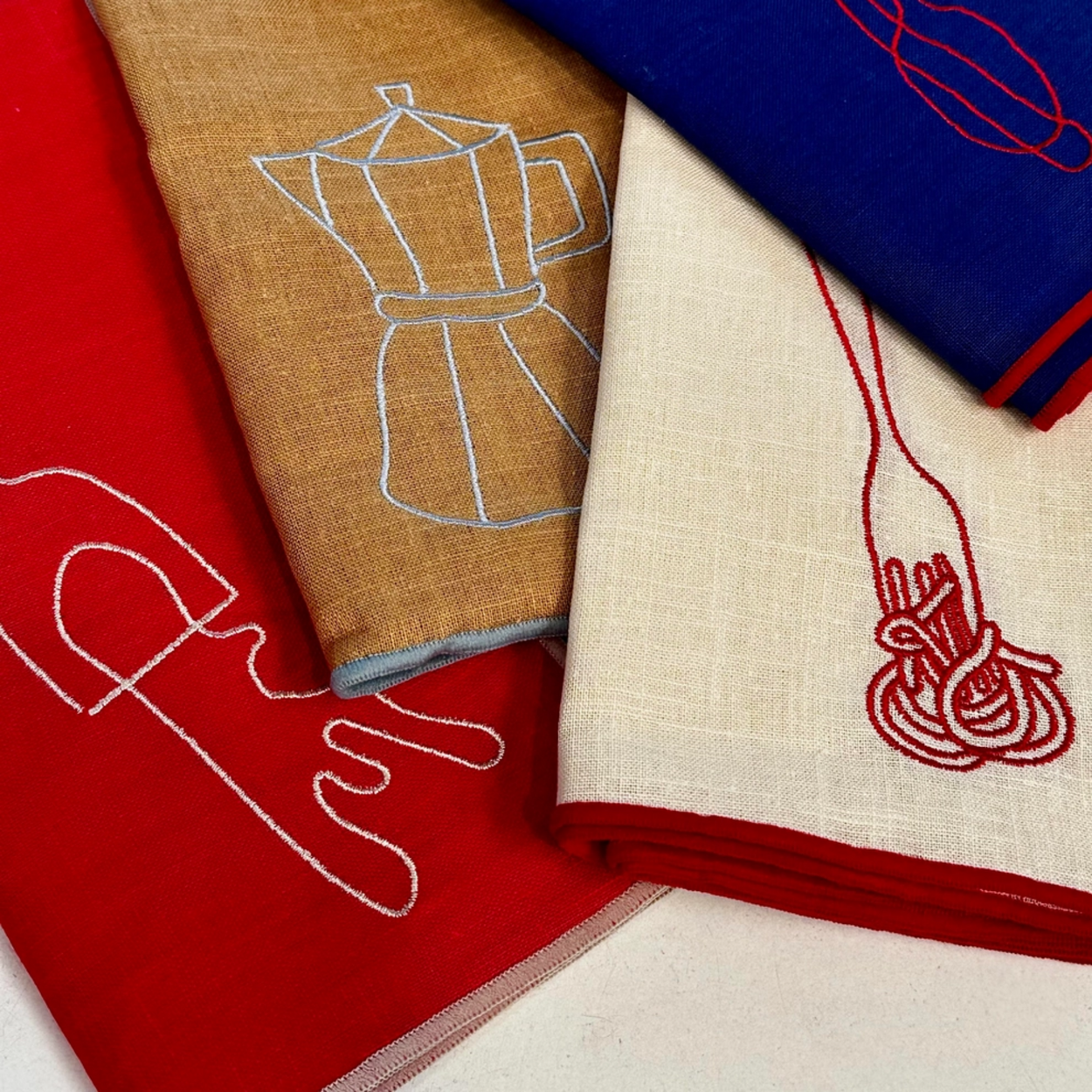 Four folded linen napkins in red, tan, blue, and cream, each featuring a minimalist embroidered kitchen utensil or food outline in contrasting thread.