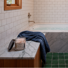 A modern bathroom interior with white subway tile walls, a freestanding bathtub over a dark marble floor, wood cabinetry, a window. A jar candle and folded navy towel sit on a marble-topped counter in the foreground.