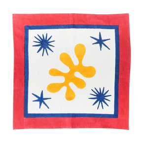 A square textile napkin, featuring a bold abstract design. The fabric has a solid red border, a white center, a thick blue inner border, a large yellow-orange splatter motif in the middle, and three blue starburst accents in the corners.
