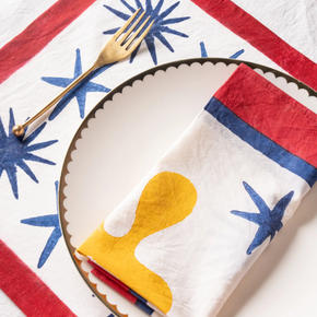 A folded white napkin with a yellow abstract shape and blue starburst motifs, styled on a scalloped white plate with red and blue stripes. A gold fork rests on the napkin over a matching striped placemat.