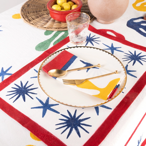 A styled table setting featuring a white tablecloth with a bold retro pattern of red borders, blue starbursts, and yellow abstract shapes. A matching red and white placemat with a gold-trimmed border is laid under a white plate with gold cutlery.